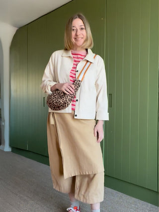 Woman standing in front of a green wall wearing a beige jacket, pink striped shirt, and beige skirt.