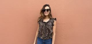 A woman wearing a dark floral ruffled blouse and blue jeans, standing against a textured terracotta wall.