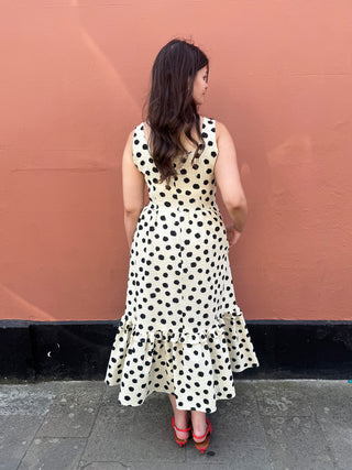 A woman stands with her back to the camera wearing a sleeveless cream dress with black polka dots and a ruffled hem, paired with red heels, against a textured terracotta wall.