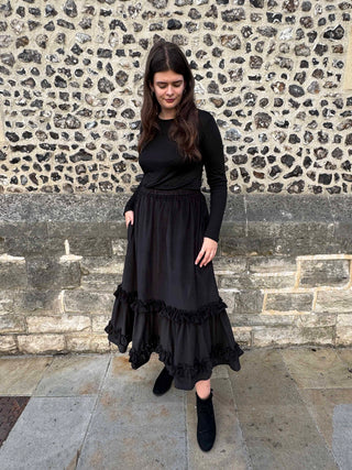 A woman wearing a black long-sleeve top and a long black ruffled skirt, standing outdoors in front of a stone wall.