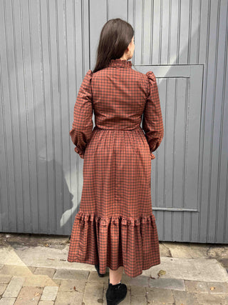 A woman viewed from behind wearing a rust-orange and black gingham midi dress with puff sleeves, a ruffled high neck, and a tiered ruffle hem, standing in front of a gray wooden door.