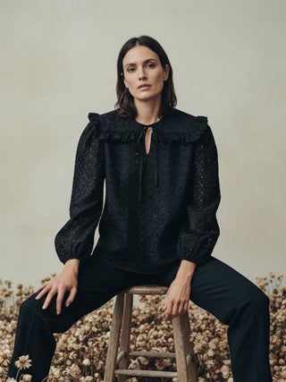 Woman sitting on a wooden stool wearing a black blouse with ruffled collar and long sleeves.