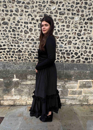A woman wearing a long-sleeved black midi dress with a ruffled hem, standing in front of a textured stone wall.