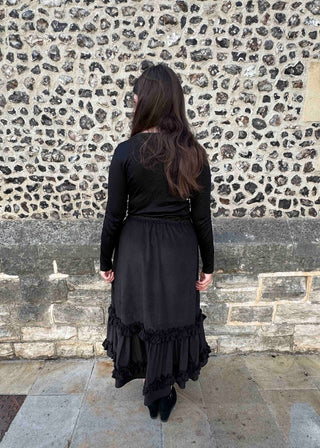 A woman viewed from behind wearing a long-sleeved black dress with a ruffled hem, standing in front of a textured stone wall.