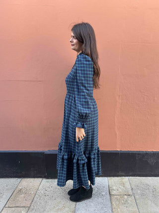 A woman wearing a dark blue and black plaid midi dress with long sleeves and ruffled tiers, standing in profile against a textured terracotta wall.