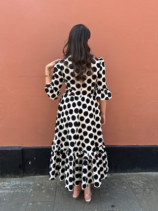 A woman viewed from behind wearing a black and white polka dot midi dress with ruffled sleeves and a ruffled hem, standing against a solid terracotta-colored wall.