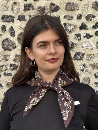 A woman with dark brown hair wearing a dark brown paisley neck scarf with gold and white patterns, tied around her collar, and a black long-sleeve top, standing in front of a textured stone wall.