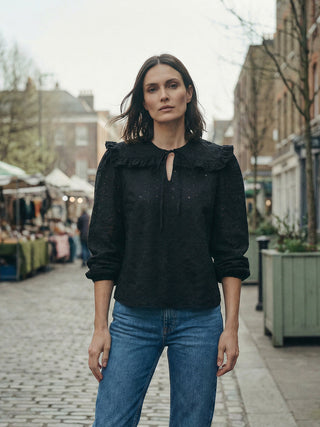 Woman wearing a black blouse with ruffled details and blue jeans standing on a street.