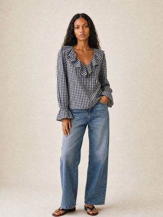 A woman modeling a black and white gingham long-sleeve blouse with a ruffled v-neckline, paired with wide-leg blue jeans and brown sandals, against a neutral beige studio background.