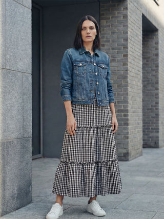 Woman wearing a denim jacket and checkered dress standing against a gray building.