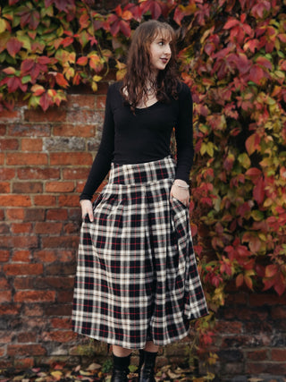 Woman wearing a black top and plaid skirt standing against a brick wall with autumn foliage.