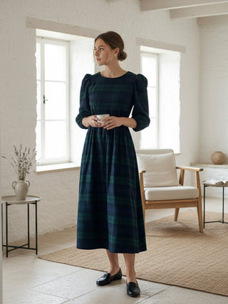 Woman in a dark green plaid dress standing in a bright room with white walls and wooden furniture.