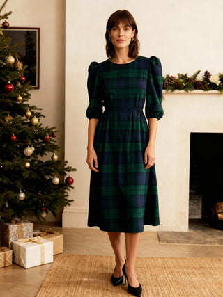 Woman in a green plaid dress standing in a festive room with a Christmas tree and fireplace.