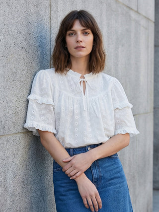 Woman wearing a white blouse with ruffled sleeves and blue jeans against a gray wall.