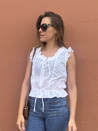 A woman wearing a white eyelet lace sleeveless top with ruffle details and a tie at the neckline, paired with blue jeans, standing against a textured terracotta wall.