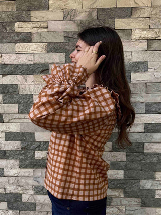 A woman wearing a rust-orange and brown gingham long-sleeve blouse with dramatic ruffled shoulders, posing in front of a gray stone tile wall.