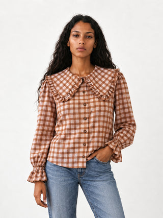 A woman modeling a brown and beige gingham long-sleeve blouse with a large ruffled Peter Pan collar and button front, paired with blue jeans, against a plain white studio background.