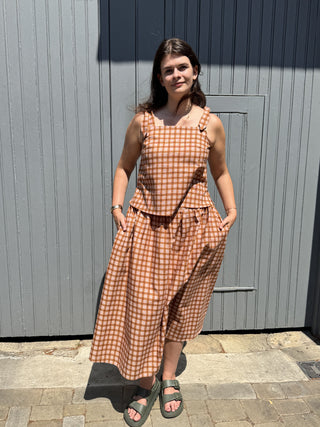 A woman wearing a rust-orange and white gingham sleeveless jumpsuit with shoulder straps and side pockets, standing outdoors in front of a gray paneled wall.