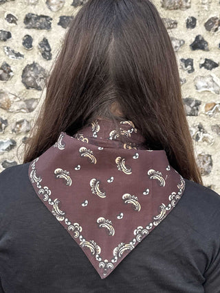 A person viewed from behind wearing a dark brown bandana with a white and gold paisley pattern around their neck, set against a rustic stone wall background.
