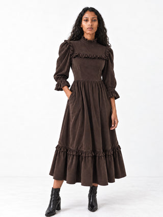 A woman modeling a dark brown midi dress with long ruffled sleeves, a high ruffled neckline, and tiered ruffles at the waist and hem, paired with black ankle boots against a white studio background.