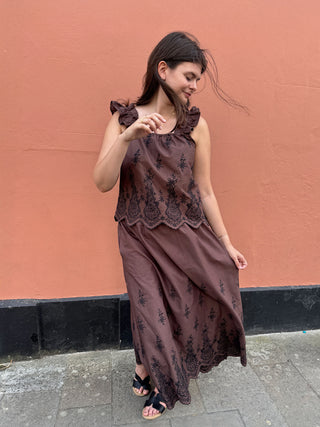 A woman wearing a brown sleeveless skirt and top with black floral embroidery and lace trim, standing against a textured terracotta wall.