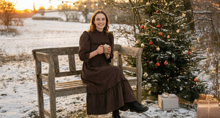 Woman in a brown dress sitting on a bench next to a decorated Christmas tree in a snowy landscape.