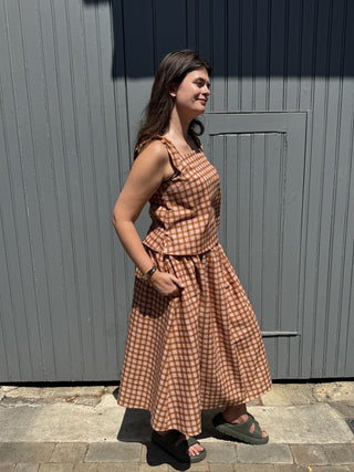 A woman wearing a brown and beige gingham midi dress with a peplum waist, standing outdoors in front of a gray corrugated metal wall.