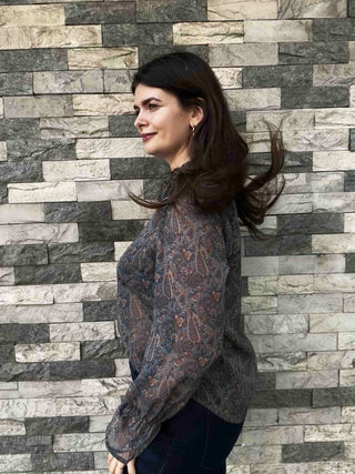 A woman with long dark brown hair is shown in profile, wearing a sheer, long-sleeve lace blouse with an intricate paisley pattern in muted brown, gray, and rust tones. She is standing in front of a gray and white stone brick wall.