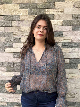 A woman with long dark brown hair stands in front of a stacked stone wall, wearing a sheer long-sleeve blouse with a blue, gray, and orange paisley pattern, paired with dark blue jeans.