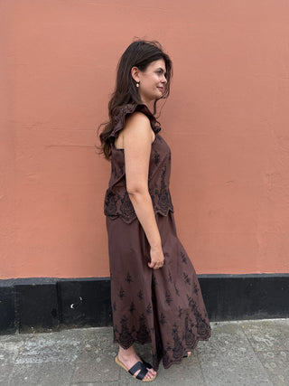 A woman wearing a sleeveless brown skirt and top with lace applique details, standing in profile against a terracotta-colored wall.
