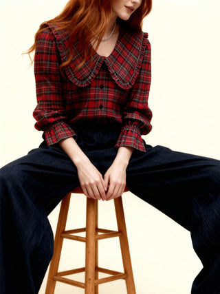Person wearing a red plaid shirt and dark pants sitting on a wooden stool against a plain background