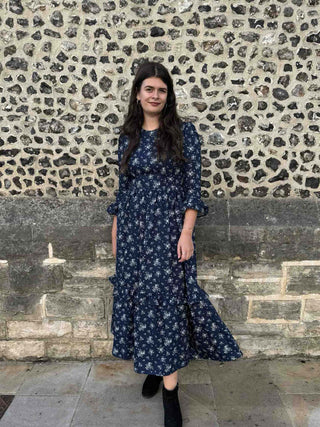 A woman wearing a navy blue midi dress with a white floral pattern, standing in front of a rustic stone wall.