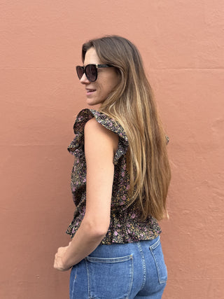 A woman with long light brown hair and sunglasses, wearing a sleeveless ruffled floral top and blue jeans, standing against a textured terracotta wall.
