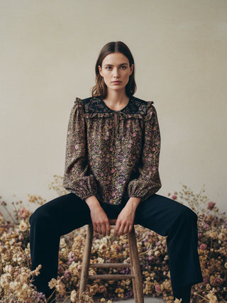 Woman sitting on a stool in front of a floral background wearing a patterned blouse and dark pants.