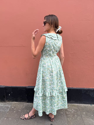 A woman wearing a sleeveless light green floral midi dress with a ruffled collar, standing in profile against a textured terracotta wall.