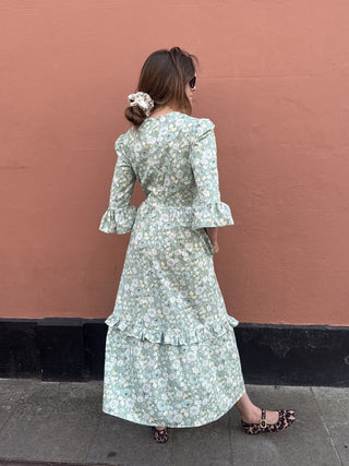 A woman stands with her back to the camera wearing a light sage green and white floral print midi dress with ruffled details, paired with leopard print flat shoes, set against a solid terracotta-colored wall.
