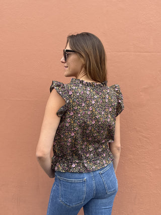 A woman wearing a dark floral ruffle-sleeve top and blue jeans, photographed from the back against a textured terracotta wall.