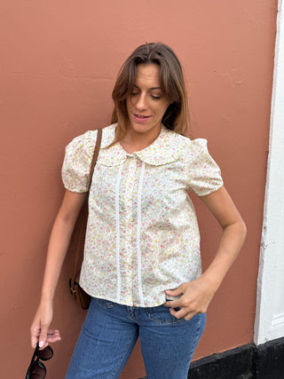 A woman wearing a short-sleeve cream-colored blouse with a delicate multicolor floral print and a Peter Pan collar, paired with blue jeans, standing against a terracotta-colored wall.
