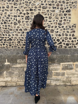 Back of A woman wearing a navy blue midi dress with a white floral pattern, standing in front of a rustic stone wall.