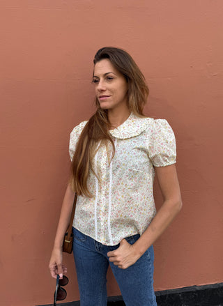 A woman wearing a short-sleeve cream-colored blouse with a delicate multicolor floral print and a ruffled Peter Pan collar, paired with blue jeans, standing against a solid terracotta-colored wall.