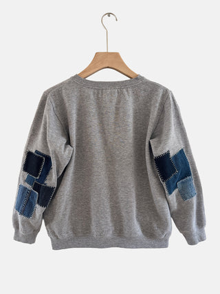 Gray sweater with denim patch details on a hanger against a white background