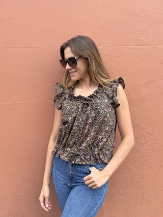 A woman wearing a dark floral ruffle-sleeve blouse and blue jeans stands against a textured terracotta wall.