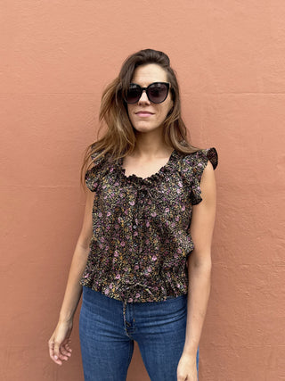 A woman wearing a dark floral ruffled blouse and blue jeans, standing against a textured terracotta wall.