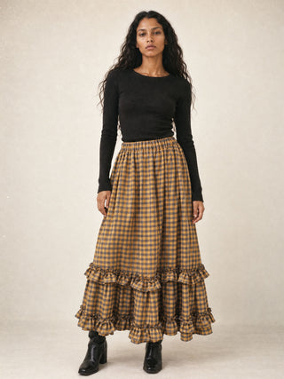 A woman stands against a plain beige studio background wearing a long black long-sleeve top and a brown and tan gingham maxi skirt with tiered ruffles at the hem, paired with black ankle boots.