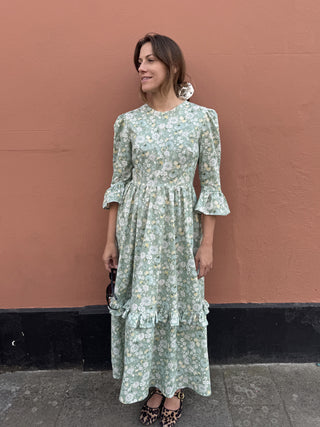 A woman wearing a light green and white floral midi dress with three-quarter ruffled sleeves, standing against a terracotta-colored wall.
