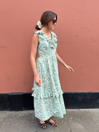 A woman wearing a light green floral tiered midi dress with ruffles, standing against a textured terracotta wall.