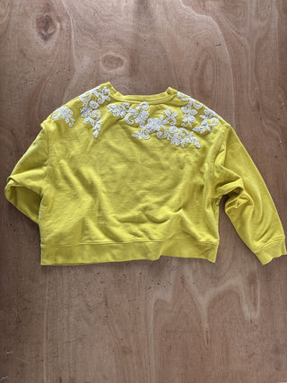 A yellow cropped sweatshirt with white floral embroidery across the shoulders, laid flat on a wooden surface.