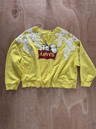 A yellow Levi's crewneck sweatshirt featuring a Snoopy and Charlie graphic on the chest, white floral embroidery on the shoulders, and a distressed raw-edge hem, laid flat on a wooden surface.