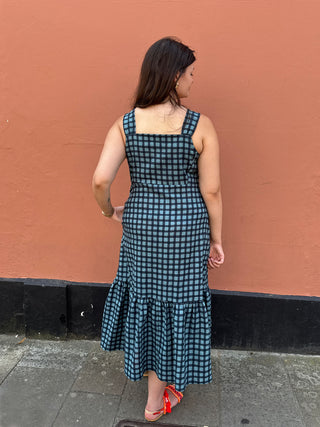 A woman stands with her back to the camera wearing a sleeveless dark blue and black gingham midi dress with a ruffled hem, set against a terracotta-colored wall.