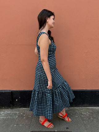 A woman wearing a sleeveless black and white gingham tiered maxi dress and red sandals, standing in profile against a textured terracotta wall.